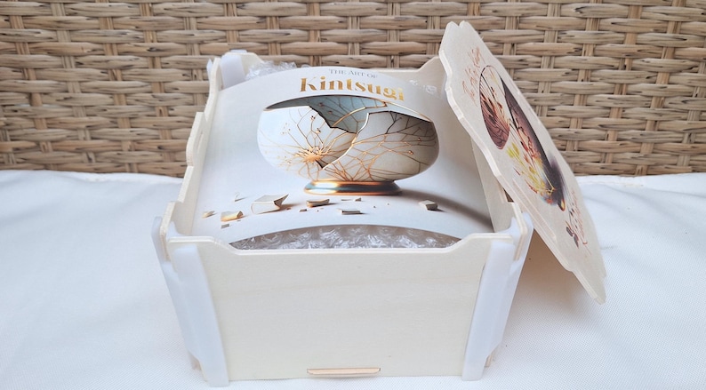 May include: A white wooden box with a lid. The lid has a printed image of a broken bowl with gold accents. The box is labeled "The Art of Kintsugi".