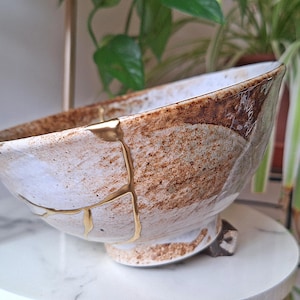 Può includere: Una ciotola in ceramica bianca con accenti marroni e dorati. La ciotola ha una glassa craquelé ed è decorata con foglia d'oro. La ciotola è appoggiata su una superficie bianca.