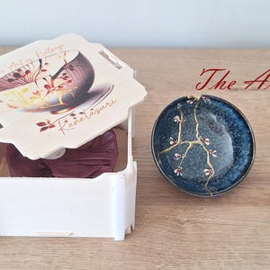 May include: A white box with a lid featuring a floral design and the text "The Art of Kintsugi" and "Kanelasuuri". The lid is open and a small blue and gold bowl with a crack repaired with gold is visible.