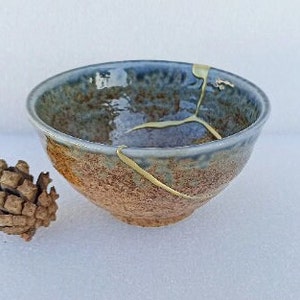 May include: A small, blue and brown ceramic bowl with a crack filled with gold paint. The bowl has a rustic, handmade look.