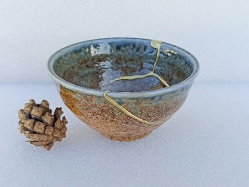May include: A small, blue and brown ceramic bowl with a crack filled with gold paint. The bowl has a rustic, handmade look.