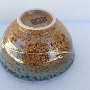 May include: A brown and blue ceramic bowl with a gold rim and a white base. The bowl has a textured surface and a unique glaze. The bottom of the bowl has a small, black stamp.
