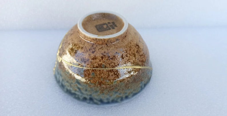 May include: A brown and blue ceramic bowl with a gold rim and a white base. The bowl has a textured surface and a unique glaze. The bottom of the bowl has a small, black stamp.