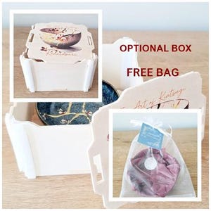 May include: A wooden box with a white lid and a white interior. The lid has a printed image of a teacup with a floral design. The box is sitting on a wooden surface. The text on the lid reads "Kintsugi". The box is being used to package a small, pink, fabric-wrapped bowl. The bowl is sitting inside a clear, organza bag. The bag has a small, white tag attached to it. The tag has the text "The Art of Kintsugi" printed on it. The box is an optional purchase, and the bag is included for free.
