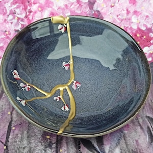 May include: A dark blue ceramic bowl with a gold-colored kintsugi repair. The bowl features a floral design with pink and white blossoms. The background shows a pink floral painting.