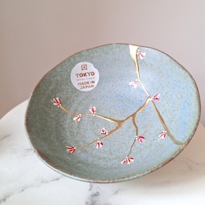 May include: A round, light blue and green ceramic bowl with gold-colored kintsugi-style repairs and red floral accents. The bowl has a sticker that reads "TOKYO DESIGN STUDIO MADE IN JAPAN".
