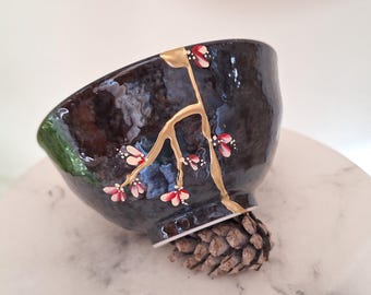 May include: A dark, glossy, textured ceramic bowl featuring a gold-colored vein design that resembles kintsugi. The bowl is decorated with small, hand-painted pink and white floral blossoms along the gold lines, creating a delicate contrast.