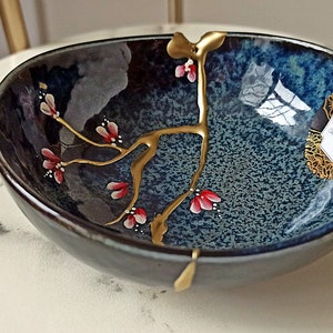 Puede incluir: Un peque&ntilde;o cuenco de cer&aacute;mica azul oscuro con un dise&ntilde;o floral dorado y rojo. El cuenco est&aacute; sobre una superficie de m&aacute;rmol blanco.