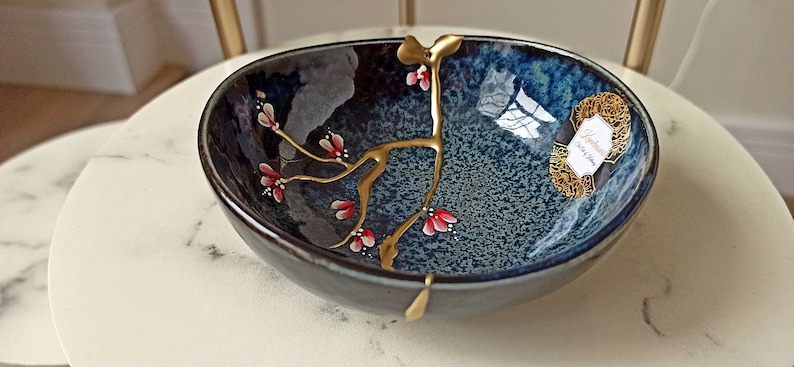 Puede incluir: Un peque&ntilde;o cuenco de cer&aacute;mica azul oscuro con un dise&ntilde;o floral dorado y rojo. El cuenco est&aacute; sobre una superficie de m&aacute;rmol blanco.