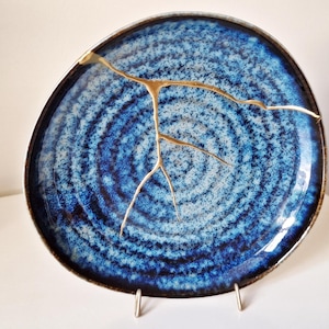 Könnte beinhalten: Ein blauer und weißer Keramikteller mit goldfarbenem Craquelé-Design. Der Teller steht auf einem Ständer und hat eine runde Form.