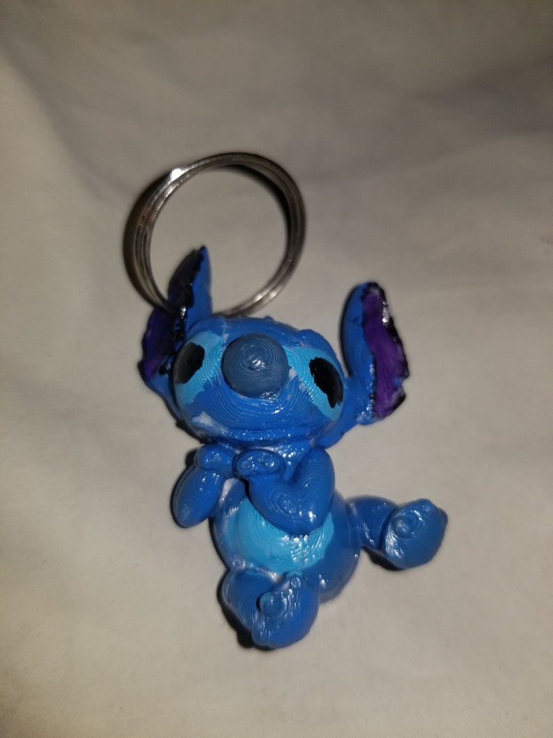 May include: Blue Stitch keychain with a silver ring. The Stitch figure is 3D printed and has a blue, purple, and white paint job.