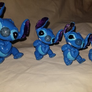 May include: Four blue Stitch figurines of varying sizes, with purple ears and white accents. The figurines are made of a smooth, painted material and are posed in a sitting position with their arms crossed.