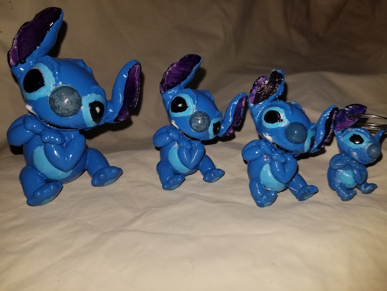 May include: Four blue Stitch figurines of varying sizes, with purple ears and white accents. The figurines are made of a smooth, painted material and are posed in a sitting position with their arms crossed.