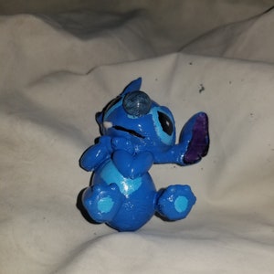 May include: Blue Stitch figurine with a white and blue heart on its chest. The figurine is lying on its back with its arms crossed and its legs bent.