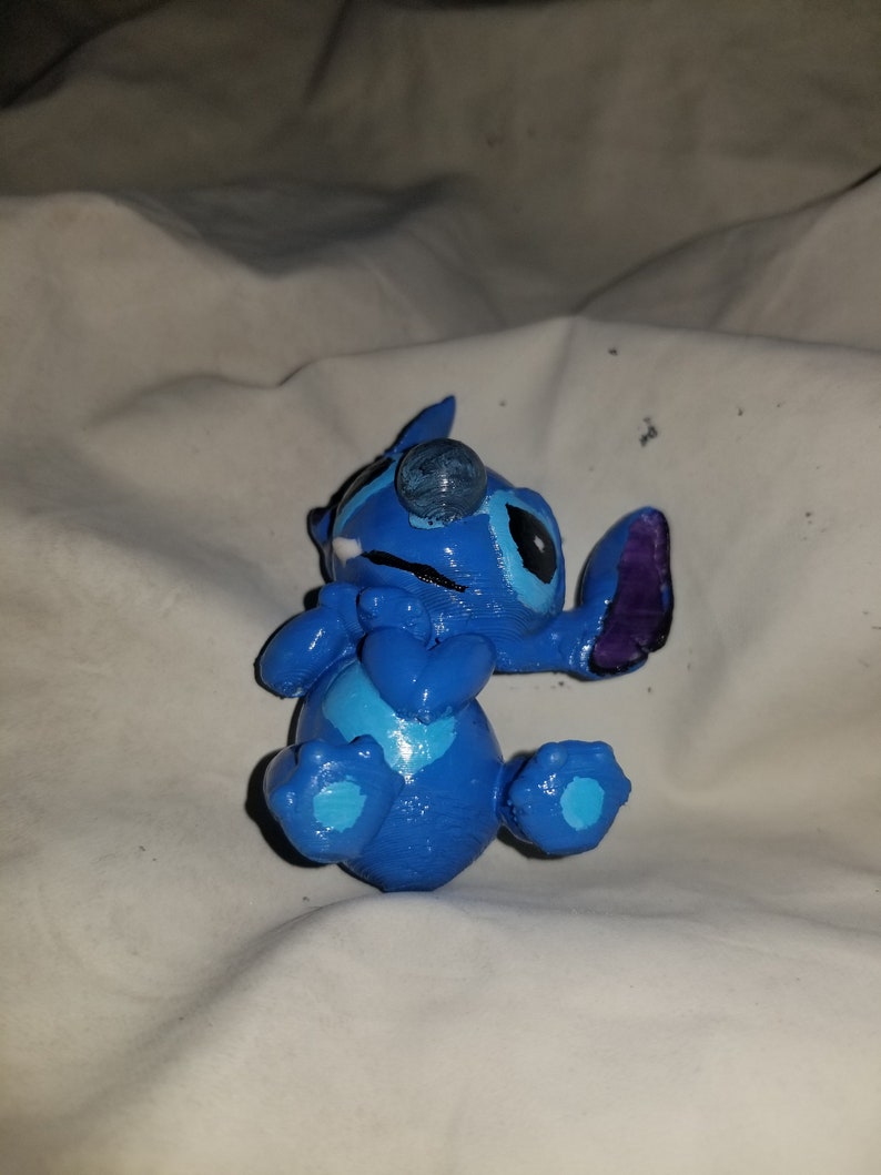 May include: Blue Stitch figurine with a white and blue heart on its chest. The figurine is lying on its back with its arms crossed and its legs bent.