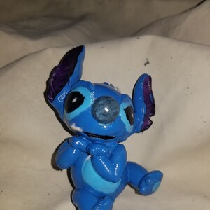 May include: A blue and purple Stitch figurine with a white belly and blue eyes. The figurine is sitting with its hands clasped in front of it.