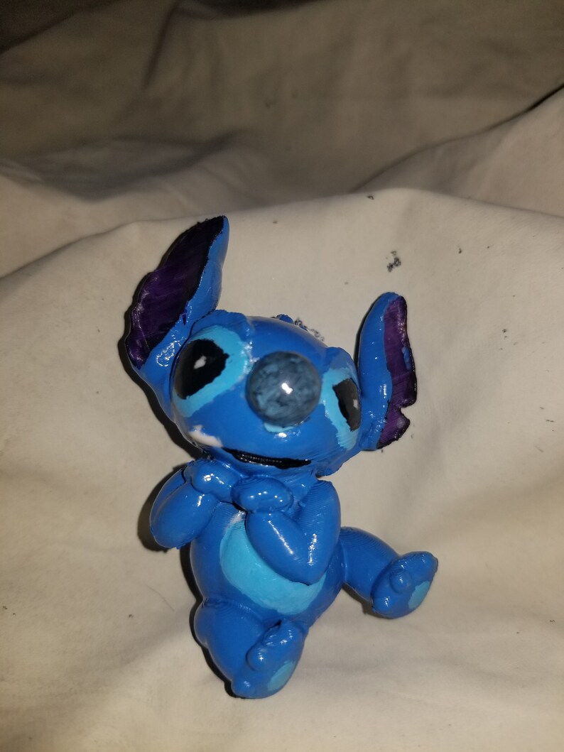 May include: A blue and purple Stitch figurine with a white belly and blue eyes. The figurine is sitting with its hands clasped in front of it.