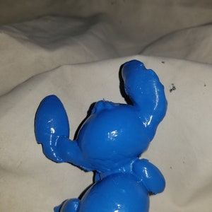 May include: A blue plastic figurine of Stitch from the Disney movie Lilo & Stitch. The figurine is posed with its arms raised and its head tilted back.