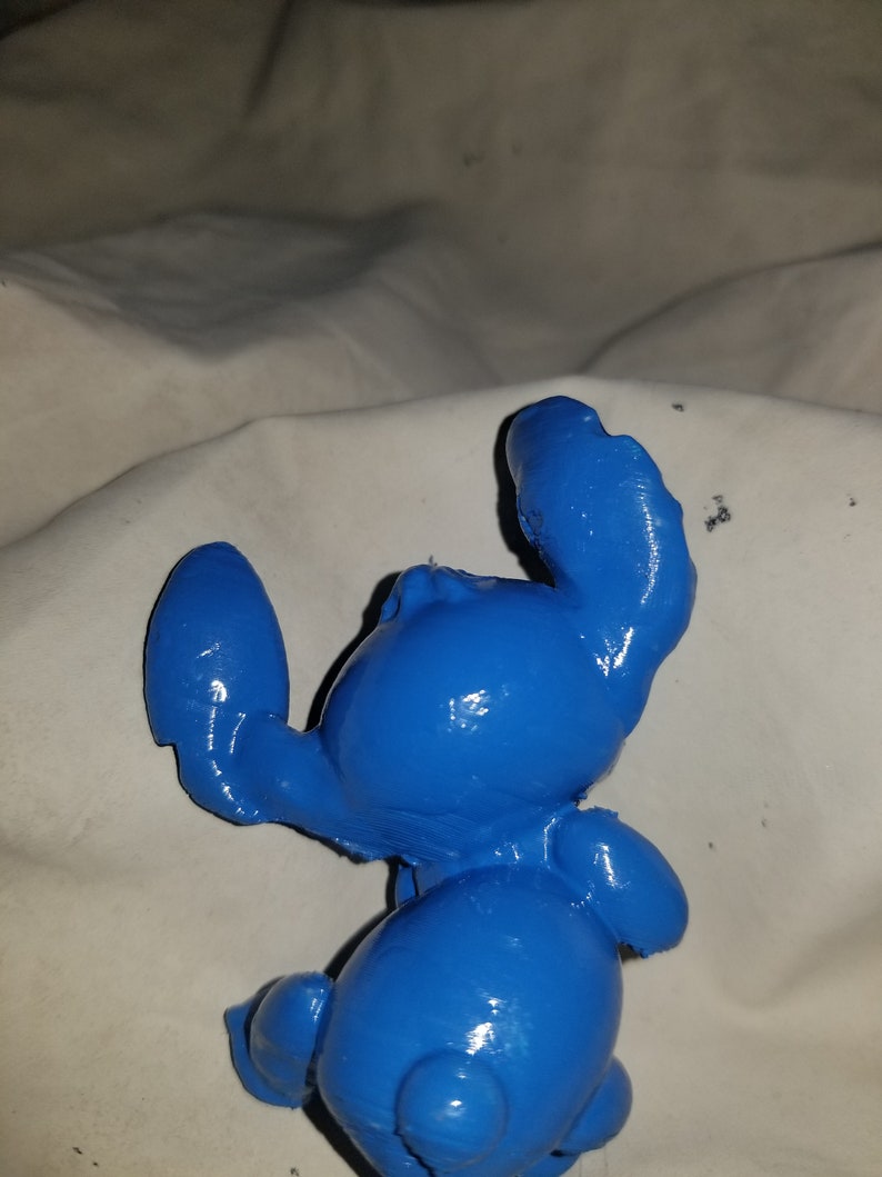 May include: A blue plastic figurine of Stitch from the Disney movie Lilo & Stitch. The figurine is posed with its arms raised and its head tilted back.