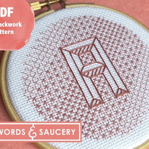 Könnte beinhalten: Eine Nahaufnahme eines fertigen Stickrahmens mit einem geometrischen Muster in Rosa und Weiß. Das Muster ist ein Blackwork-Design des Buchstabens "A" mit einem sich wiederholenden Sternmuster darum herum. Der Rahmen ist aus Holz und hat eine natürliche Oberfläche. Der Text "WORDS & SAUCERY" ist am unteren Rand des Rahmens sichtbar. Der Text "PDF Blackwork Pattern" ist in der oberen linken Ecke des Bildes sichtbar.