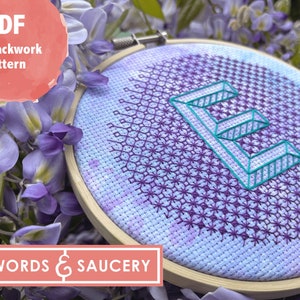 Könnte beinhalten: Nahaufnahme eines fertigen Stickrahmens mit einem lila und blauen Farbverlauf im Hintergrund. Der Buchstabe "E" ist in Türkis und Weiß gestickt, mit einem schwarzen Stickmuster im Hintergrund. Der Rahmen ist von lila Glyzinienblüten umgeben.