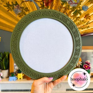 Peut inclure: Un cercle à broder vert olive avec un motif de nœuds celtiques sur le bord. Le cercle contient du tissu blanc, idéal pour le point de croix ou la broderie. Le cercle est tenu dans la main d'une personne.