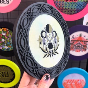 May include: A round, framed cross-stitch with a dark grey Celtic knot frame. The cross-stitch design features a black pot, two houses, and a cannabis leaf. Other colourful cross-stitch pieces are visible in the background.