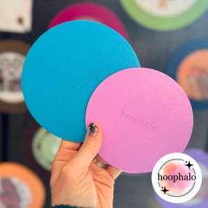 Puede incluir: Dos alfombrillas redondas, de color liso, en azul brillante y rosa, sostenidas por una mano. Las alfombrillas tienen la palabra "hoophalo" en relieve. En el fondo se ven más artículos similares, lo que sugiere una colección de productos relacionados.