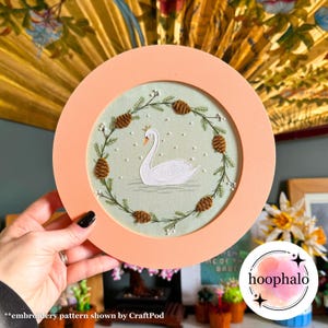 May include: A white swan embroidered on a light green fabric with a brown pine cone wreath. The embroidery is framed in a light pink round frame.