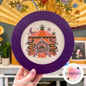 Könnte beinhalten: Eine lila gerahmte Kreuzstich-Stickerei eines rosa und orangefarbenen Hauses mit einem grünen Dach und einem Mond und Sternen am Himmel. Die Stickerei trägt den Titel "Hoop Halo" und stammt von Innocent Bones.