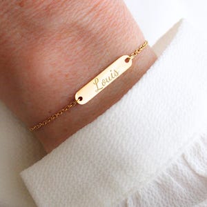 Peut inclure: Bracelet doré avec une plaque rectangulaire gravée du nom "Louis". Le bracelet est attaché à une chaîne délicate. Le bracelet est porté sur un poignet, contre une chemise blanche.