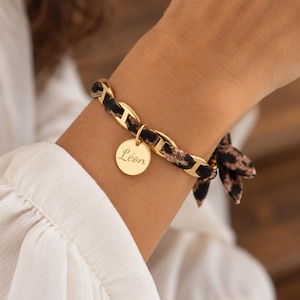 Peut inclure: Un bracelet à maillons dorés entrelacé d'un tissu à motif léopard. Un pendentif rond doré est suspendu au bracelet, gravé du prénom Léon en écriture cursive. Le bracelet se termine par un nœud en tissu assorti au motif léopard.