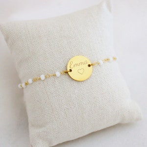 May include: A gold bracelet with a round charm that says "Emma" with a small heart engraved on it. The bracelet is made of a delicate chain with small white beads.