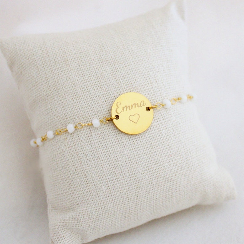 May include: A gold bracelet with a round charm that says "Emma" with a small heart engraved on it. The bracelet is made of a delicate chain with small white beads.