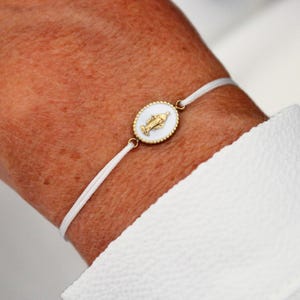 Peut inclure: Bracelet en cordon blanc orné d'un pendentif ovale doré avec un centre blanc et une figure religieuse. Le pendentif est entouré d'une bordure perlée dorée. Le bracelet est porté au poignet, avec une manche de chemise blanche visible.