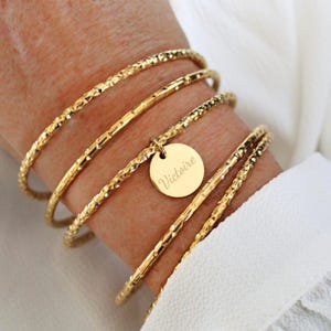 Peut inclure: Un ensemble de cinq bracelets joncs dorés, dont quatre présentent une surface texturée et facettée. Une breloque ronde et lisse, également dorée, est suspendue au centre, gravée du prénom Victoire en écriture cursive.