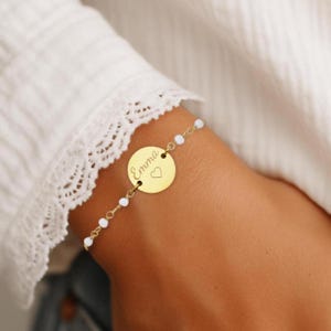 May include: A gold bracelet with a round charm that says "Emma" with a small heart engraved on it. The bracelet is made of small white beads and a gold chain.