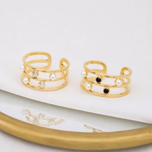 Peut inclure: Deux bagues ouvertes dorées au design à trois anneaux. La bague de gauche est ornée de perles blanches et de cristaux transparents. La bague de droite présente des perles blanches et des accents circulaires noirs. Ouvertes pour le réglage.