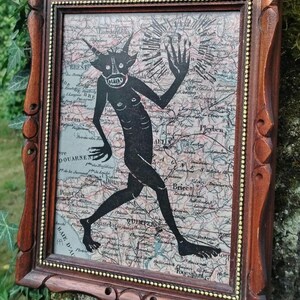 Peut inclure: Une œuvre encadrée représentant une silhouette noire d'une figure cornue sur un fond de carte ancienne. La figure sombre est en mouvement, un bras levé. Le cadre est orné, en bois foncé avec un bord intérieur doré.