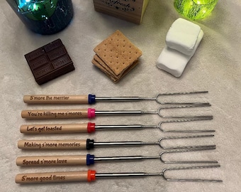 May include: A collection of s'mores roasting forks with wooden handles and stainless steel prongs. Each handle has a different colored accent and a unique engraved phrase, including "S'more the merrier" and "S'more good times". The image also shows graham crackers, marshmallows, and chocolate.