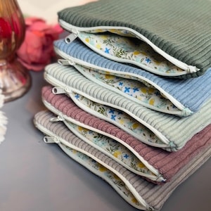 May include: A stack of six zippered pouches with different colored corduroy exteriors and a floral print lining. The pouches are stacked in order from green, blue, gray, pink, gray, and beige.