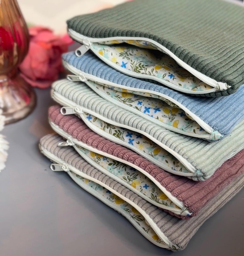 May include: A stack of six zippered pouches with different colored corduroy exteriors and a floral print lining. The pouches are stacked in order from green, blue, gray, pink, gray, and beige.