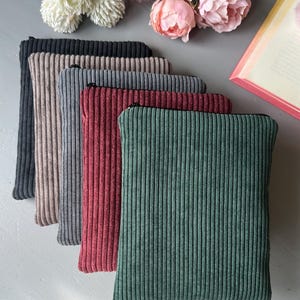 Könnte beinhalten: Sechs Cord-Reißverschlusstaschen in verschiedenen Farben: Schwarz, Beige, Grau, Burgunderrot und Grün. Die Taschen sind in einer Reihe angeordnet und haben einen schwarzen Reißverschluss. Im Hintergrund befinden sich Blumen und eine Glasvase.
