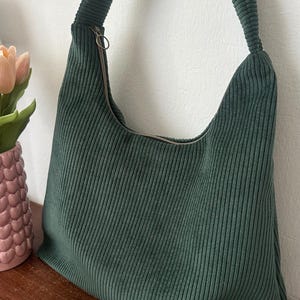 Könnte beinhalten: Grüne Cord-Umhängetasche mit Reißverschluss. Die Tasche hat einen breiten Riemen und ein schlichtes Design. Die Tasche liegt auf einer Holzoberfläche. Maße in Zentimetern.
