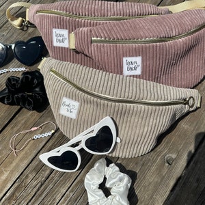 Könnte beinhalten: Zwei braune Cord-Bauchtaschen mit goldenen Reißverschlüssen und weißen Patches, auf denen "Team Bride" und "Bride to Be" steht. Die Bauchtaschen liegen auf einer Holzoberfläche mit einer weißen Herz-Sonnenbrille, einem weißen Haargummi und einem Perlenarmband, auf dem "BRIDE" steht.