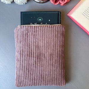 May include: A pink corduroy book sleeve with a zipper closure. The book sleeve is holding a book with a dark blue cover and a green eye design. The text on the book cover reads "Der Herr der Eyvindens".