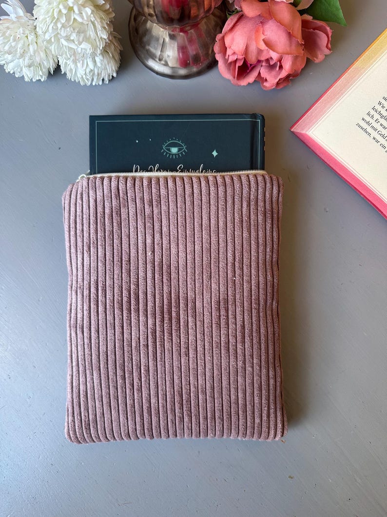 May include: A pink corduroy book sleeve with a zipper closure. The book sleeve is holding a book with a dark blue cover and a green eye design. The text on the book cover reads "Der Herr der Eyvindens".