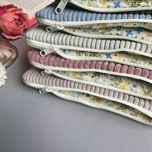May include: A stack of six zippered pouches with floral fabric lining. The pouches are made of corduroy fabric in various colors, including green, blue, gray, and pink.