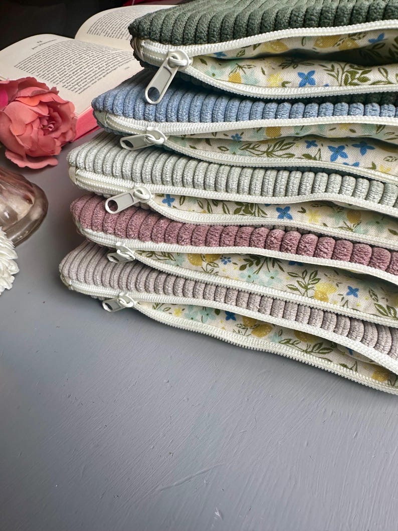 May include: A stack of six zippered pouches with floral fabric lining. The pouches are made of corduroy fabric in various colors, including green, blue, gray, and pink.