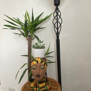 Può includere: Una testa di manichino in bianco e nero con un turbante e una sciarpa a motivi gialli, verdi e neri. La testa è seduta su uno sgabello di vimini con una pianta verde in un vaso bianco dietro di essa. Una lampada da terra nera con un paralume bianco è sullo sfondo.